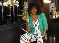 A young Black woman in a green blazer on a bicycle holding a pineapple. 