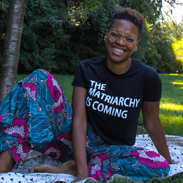 A young Black woman sitting on the grass wearing a shirt that says "The matriarchy is coming"